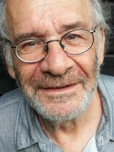 Zbliżenie twarzy starszego mężczyzny z siwymi włosami, krótką brodą i okrągłymi okularami. Uśmiecha się delikatnie, patrząc w obiektyw. Ubrany jest w jasnoszarą koszulę, a zdjęcie ma naturalny, kameralny charakter.