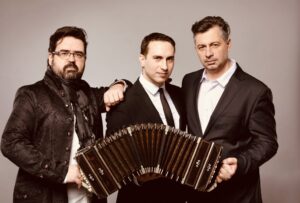Trzech muzyków pozujących do zdjęcia studyjnego; centralnie widoczny jest bandoneon trzymany przez jednego z nich.