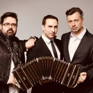 Trzech muzyków pozujących do zdjęcia studyjnego; centralnie widoczny jest bandoneon trzymany przez jednego z nich.