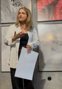 Kobieta stoi w galerii przed ścianą z oprawionymi pracami i przemawia do publiczności. Ubrana jest w białą koszulę narzuconą na ciemny strój, w jednej ręce trzyma kartkę, a drugą gestykuluje. W tle widoczne są plakaty i grafiki wystawy, a wnętrze ma drewnianą podłogę i industrialny charakter