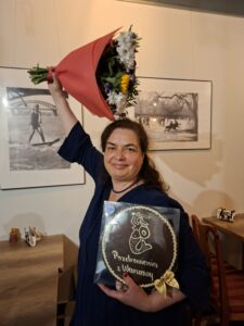 Kobieta w granatowej sukience stoi w kawiarni lub galerii, uśmiechając się do obiektywu. W uniesionej ręce trzyma duży bukiet kolorowych kwiatów, a w drugiej okrągły piernik z napisem „Pozdrowienia z Warszawy” i wizerunkiem syrenki. W tle widoczne są stoliki oraz czarno-białe fotografie wiszące na ścianie.
