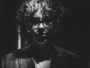 Czarno-biały portret osoby z krótkimi, kręconymi włosami i dużymi ciemnymi okularami. Ubrana jest w marynarkę w kratę i jasną koszulkę. Twarz częściowo oświetla punktowe światło, a ciemne tło nadaje fotografii wyrazisty, kontrastowy i artystyczny charakter