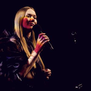 Katarzyna Dąbrowska śpiewa do mikrofonu na ciemnej scenie, oświetlona punktowym światłem podkreślającym jej twarz i czarny sceniczny strój.